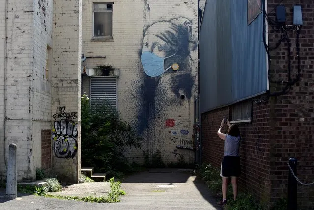 A person takes a picture of a piece of Banksy street art titled “The Girl with the Pierced Eardrum”, now adorned with a protective face mask, at Albion Dock in Bristol, Britain on April 22, 2020. It depicts a take on Vermeer’s famous Girl with a Pearl Earring, replacing the earring with an outdoor security alarm. The art first appeared on the side of a building in Albion Docks in Hanover Place, in Bristol's Harbourside in October 2014. (Photo by Rebecca Naden/Reuters)
