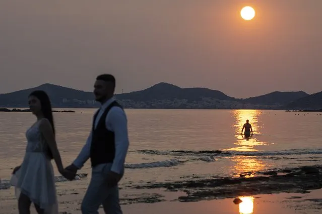 A newly married couple poses for photos during sunset as a man takes a dip in the water in Lagonisi southeast of Athens, Wednesday, July 12, 2023. The government has announced emergency measures for later this week, allowing workers to stay home during peak temperature hours as a heat wave is due to affect most of Greece. (Photo by Petros Giannakouris/AP Photo)