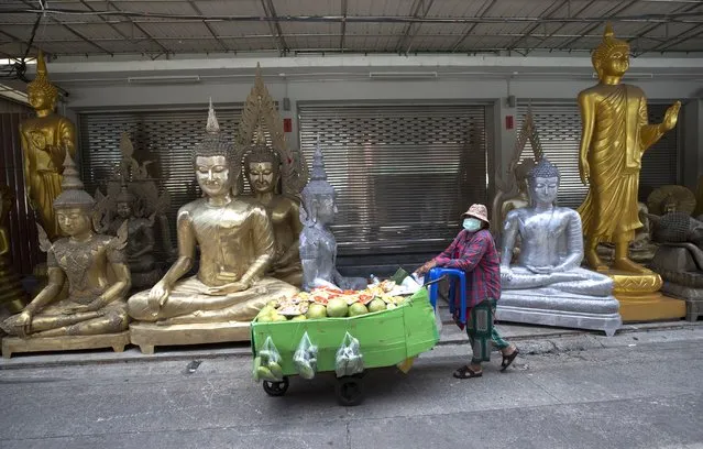 A fruit vendor wearing a protective mask to help curb the spread of the coronavirus pushes her cart past giant Buddhist statues for sale in Bangkok, Thailand, Thursday, May 27, 2021. (Photo by Sakchai Lalit/AP Photo)