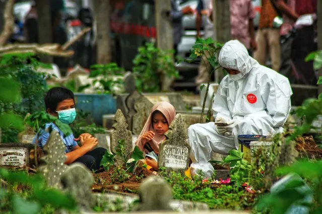 A family member in personal protective equipment (PPE) suit together with two children pray by the grave of a Covid-19 coronavirus victim who was just buried, as family members defying health protocols gather nearby at a cemetery in Lhokseumawe, Aceh on May 18, 2021. (Photo by Azwar Ipank/AFP Photo)