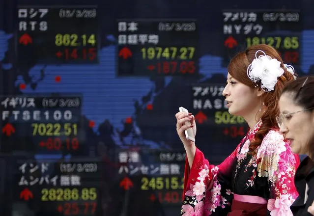A woman wearing a Hakama, or Japanese traditional Kimono, holds her mobile phone as she walks past an electronic board, showing the stock market indices of various countries, outside a brokerage in Tokyo, March 23, 2015. Asian shares started the week on a strong note on Monday after a weaker U.S. dollar helped fuel solid gains on Wall Street. (Photo by Yuya Shino/Reuters)