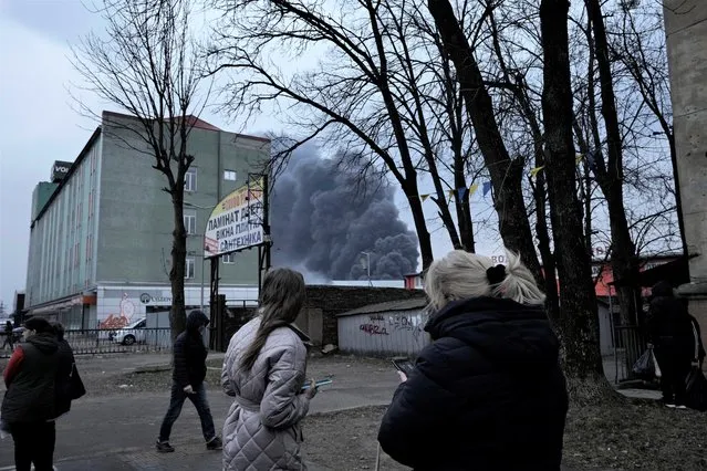 People watch smoke rising behind buildings following explosions in Lviv, western Ukraine, Saturday, March 26, 2022. (Photo by Nariman El-Mofty/AP Photo)