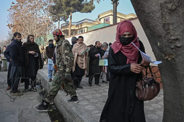 A Taliban fighter walks past people waiting to enter the passport office at a checkpoint in Kabul on December 19, 2021, after Afghanistan's Taliban authorities said they will resume issuing passports. (Photo by Mohd Rasfan/AFP Photo)