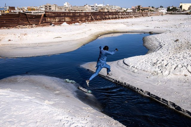 A man jumps over black water pouring out along Hann Bay in Dakar on November 22, 2023. (Photo by John Wessels/AFP Photo)