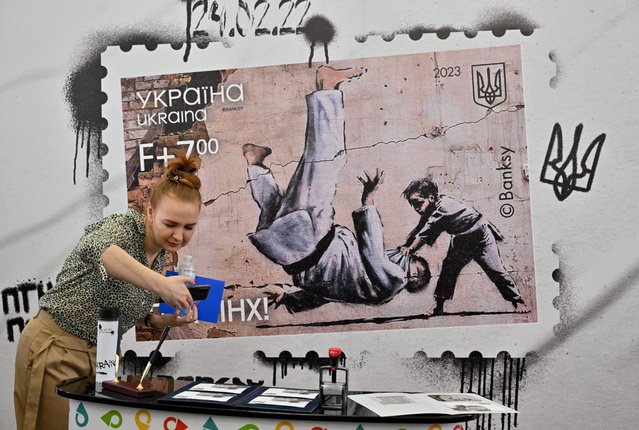 A woman takes a picture of stamps and envelopes at the main post office during the presentation of a new Ukrainian stamp with a picture of Banksy, in Kyiv on February 24, 2023, on the first anniversary of the Russian invasion of Ukraine. Residents of Kiev flocked to the main post office of the capital to get a new stamp reproducing a drawing of the famous British artist Banksy made on the wall of a destroyed building in Borodyanka. The work depicts a child judoka throwing a man also in judo clothing to the ground. On the bottom of the stamp was added the inscription “f*ck yourself Putin”, in reference to the judo practiced by the Russian president. (Photo by Sergei Supinsky/AFP Photo)