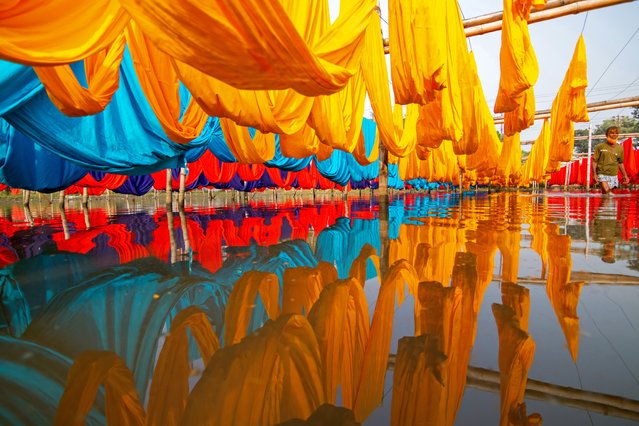 Workers hang thousands of different colorful fabrics on iron wires tied between a bamboo framework and constantly turn them so that they dry perfectly in a flooded field in Narayanganj, Bangladesh on March 16, 2022. Iron wires are used between a bamboo framework to create giant washing lines for the final part of the dying process as the fabrics are dried in the sun. Bright strands of blue, pink, orange and green-dyed cloths hang above the grassy field in a dazzling network of interlocking colors. This is the final part of the dying process after which the cloth is made into t-shirts and vests at the garment factory. (Photo by Joy Saha/ZUMA Press Wire/Rex Features/Shutterstock)