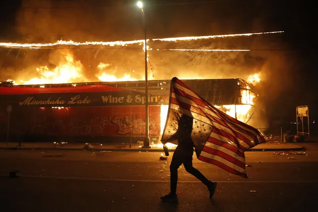 A protester carries a U.S. flag upside down, a sign of distress, next to a burning building Thursday, May 28, 2020, in Minneapolis. Protests over the death of George Floyd, a black man who died in police custody Monday, broke out in Minneapolis for a third straight night. (Photo by Julio Cortez/AP Photo)