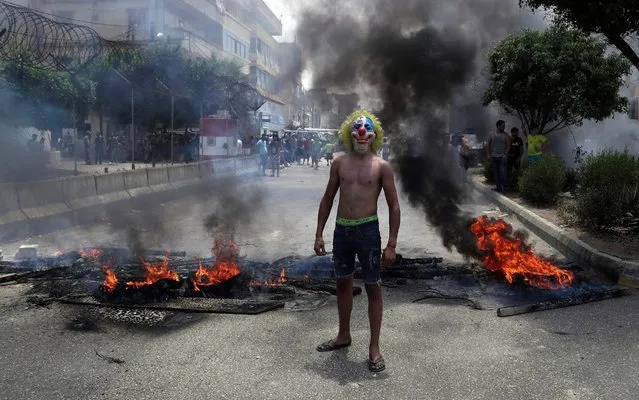 An anti-government demonstrator poses with a clown mask as others burn tires and wood to block a road in Beirut, Lebanon, Tuesday, July 14, 2020. Trash has been piling up in different parts of Lebanon recently as no deal has been reached between the government and waste management companies over payments for their employees. Most workers at the companies are foreigners and want to get paid in U.S. dollars. Lebanon is witnessing shortage in hard currency and the Lebanese pound has lost more than 80% of its value in recent months. (Photo by Bilal Hussein/AP Photo)