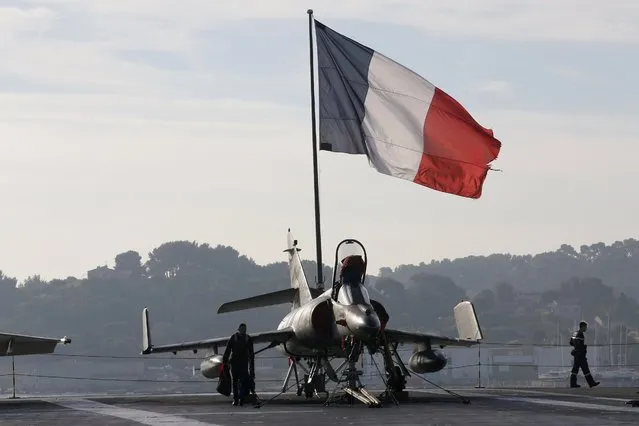 Flight deck crew work around a Super Etendard fighter jet as a French flag flies aboard the French nuclear-powered aircraft carrier Charles de Gaulle before its departure from the naval base of Toulon, France, November 18, 2015. France's Charles de Gaulle aircraft carrier will be deployed today to support operations against Islamic State in Syria and Iraq. (Photo by Jean-Paul Pelissier/Reuters)