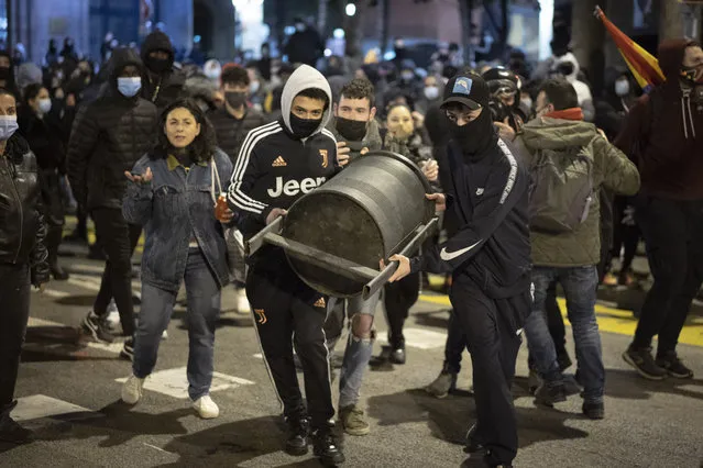 Demonstrators carry a rubbish bin to throw it agains police officers protecting a national police station during clashes following a protest condemning the arrest of rap singer Pablo Hasél in Barcelona, Spain, Sunday, February 21, 2021. Protests in support of a jailed rapper turned violent for a sixth consecutive night in Barcelona on Sunday with clashes between police and groups of mostly angry youths in the center of the Spanish city. (Photo by Emilio Morenatti/AP Photo)