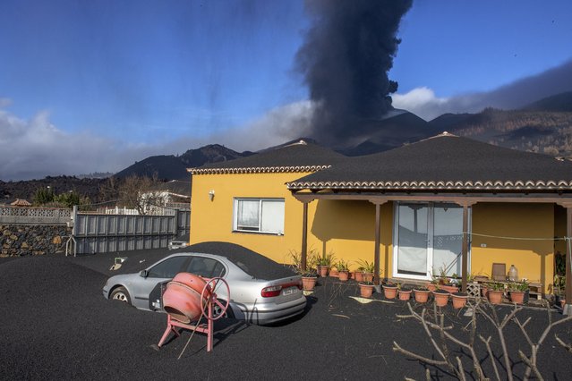 Ash covers a house, car and garden as in the background a volcano erupts on the Canary island of La Palma, Spain on Monday October 4, 2021. More earthquakes are rattling the Spanish island of La Palma, as the lava flow from an erupting volcano surged after part of the crater collapsed. Officials say they don't expect to evacuate any more people from the area, because the fiery molten rock was following the same route to the sea as earlier flows. (Photo by Saul Santos/AP Photo)