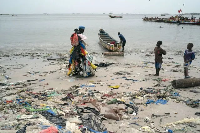 Kids look at environmental activist Modou Fall, who many simply call “Plastic Man”, while he walks on the Yarakh Beach littered by trash and plastics in Dakar, Senegal, Tuesday, November 8, 2022. As he walks, plastics dangle from his arms and legs, rustling in the wind while strands drag on the ground. On his chest, poking out from the plastics, is a sign in French that says, “No to plastic bags”. (Photo by Leo Correa/AP Photo)