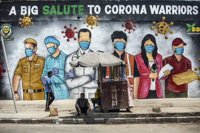 A man rests in the shade of a stall (R) while another walks past a mural showing images of frontline workers after the government eased a nationwide lockdown imposed as a preventive measure against the COVID-19 coronavirus in New Delhi on June 11, 2020. (Photo by Xavier Galiana/AFP Photo)