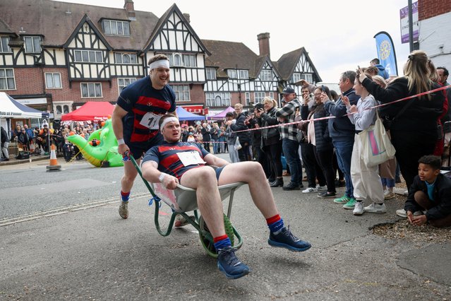 Elliot Handler and Jack O'Connor participate in the Pinner Wheelbarrow Race as part of a St. George's Day celebration in northwest London, Britain, on April 21, 2024. The race has been held for the past 61 years as part of the London suburb's St. George's Day celebrations and involves teams of two taking turns pushing each other in a wheelbarrow through the streets of Pinner while drinking beer. (Photo by Suzanne Plunkett/Reuters)