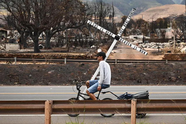 A man on a bike carries a cross in the north side of Lahaina, Hawaii, on August 16, 2023. The number of people known to have died in the horrific wildfire that levelled a Hawaiian town reached 106 on August 15, authorities said, as a makeshift morgue was expanded to deal with the tragedy. US President Joe Biden will head to fire-ravaged Hawaii on August 21 to meet with survivors and first responders still hunting for victims, the White House said on August 16. (Photo by Patrick T. Fallon/AFP Photo)