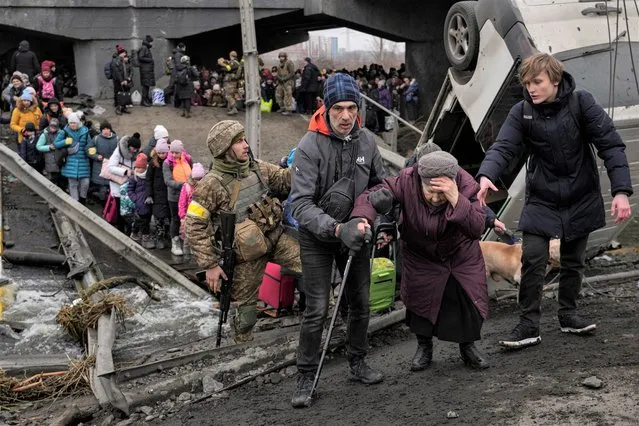 An elderly lady is assisted while crossing the Irpin river, under a bridge that was destroyed by a Russian airstrike, as civilians flee the town of Irpin, Ukraine, Saturday, March 5, 2022. What looked like a breakthrough cease-fire to evacuate residents from two cities in Ukraine quickly fell apart Saturday as Ukrainian officials said shelling had halted the work to remove civilians hours after Russia announced the deal. (Photo by Vadim Ghirda/AP Photo)