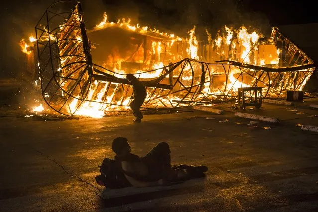 A resident, lying shirtless, keeps warm as another approaches the blazing skeleton of Juanita's Fashions R Boutique after it was burned to the ground in Ferguson, Missouri early morning November 25, 2014. Gunshots rang out and buildings burned in a Midwestern suburb after a grand jury decided on Monday not to indict a white police officer over the fatal August shooting of an unarmed black teenager, sparking a fresh wave of racially tinged violence. (Photo by Adrees Latif/Reuters)