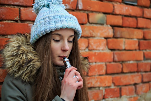 Vape teenager. Young pretty white girl in blue cap and green jacket smoking an electronic cigarette opposite brick wall on the street in the spring. Bad habit. (Photo by Aleksandr Yu/Getty Images)