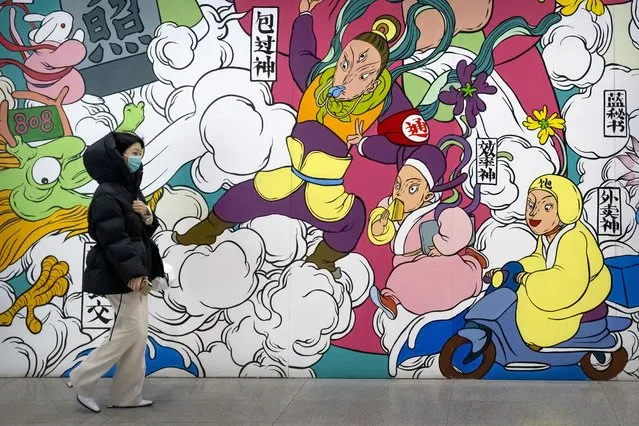 A woman wearing a face mask to protect against COVID-19 walks past a mural in the central business district in Beijing, Thursday, December 23, 2021. China ordered the lockdown of as many as 13 million people in neighborhoods and workplaces in the northern city of Xi'an following a spike in coronavirus cases, setting off panic buying just weeks before the country hosts the Winter Olympic Games. (Photo by Mark Schiefelbein/AP Photo)