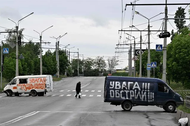 A woman carries water as she crossed street a in the Saltivka district, northern Kharkiv on May 29, 2022, amid Russian invasion of Ukraine. Inscriptions on vans reads “warning, shelling” (R) and “warning, passing by for civilians is prohibited”. (Photo by Genya Savilov/AFP Photo)