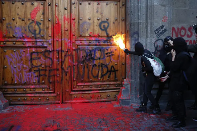 A masked, female protester sprays fire at the entrance to the National Palace, the presidential office and residence, after demonstrators covered it in fake blood and the Spanish message: “Femicide State”, in Mexico City, Friday, February 14, 2020. The demonstration against gender violence comes after last week's vicious murder of Ingrid Escamilla by her husband and controversy unleashed by the leaking of images of her body to the press, in a country where an average of 10 women are killed every day. (Photo by Ginnette Riquelme/AP Photo)