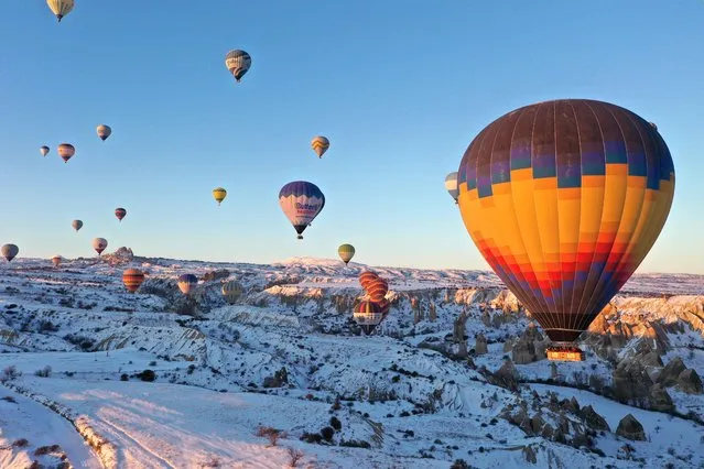 Hot-air balloons glide over snow-covered fairy chimneys in the Cappadocia region during sunrise, located in Nevsehir, Turkiye on February 11, 2022. Cappadocia is one of the vital tourism regions of Turkiye. Tourists join the hot air balloon tour and watch the natural beauties from the sky. It is considered the center of hot air ballooning globally, as it can fly 250 days a year. Cappadocia on the UNESCO World Heritage List receives tourists from various countries for 12 months. Last year, 388,833 tourists participated in hot air balloon tours in Cappadocia. (Photo by Behcet Alkan/Anadolu Agency via Getty Images)