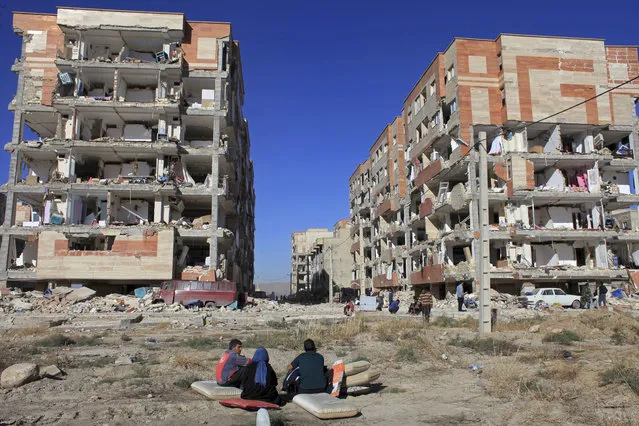 Survivors sit in front of buildings damaged by an earthquake, in Sarpol-e-Zahab, western Iran, Monday, November 13, 2017. A powerful 7.3 magnitude earthquake that struck the Iraq-Iran border region killed more than three hundred people in both countries, sent people fleeing their homes into the night and was felt as far west as the Mediterranean coast, authorities reported on Monday. (Photo by Omid Salehi/AP Photo)