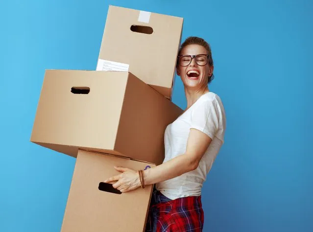 Happy young woman in white t-shirt holding pile of cardboard boxes isolated on blue. (Photo by CentralITAlliance/Getty Images/iStockphoto)