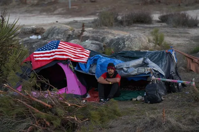 A man sits outside a tent with an American flag while he waits to be processed by US Border Patrol in Jacumba, California, USA, 28 November 2023. In recent weeks the tiny community of Jacumba, population 600, has been inundated with thousands of migrants taking temporary shelter outside in the desert due to US Border Patrol shelters being at maximum capacity. (Photo by Allison Dinner/EPA/EFE)