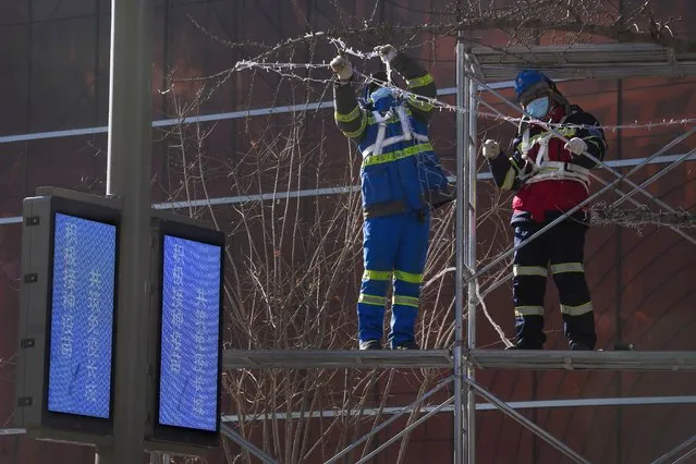 Workers wearing face masks to help protect from the coronavirus install lights on a tree for the New Year festive season near the electronic panels displaying words that reads “Actively vaccinate and build the Great Wall of Immunity” at a shopping street in Beijing, Sunday, December 26, 2021. Christmas arrived around the world Saturday amid a surge in COVID-19 infections that kept many families apart, overwhelmed hospitals and curbed religious observances as the pandemic was poised to stretch into a third year. (Photo by Andy Wong/AP Photo)