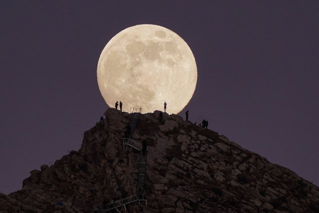 A full moon appears over a rocky hilltop as people stand silhouetted against its glow in Duhok, Iraq, on December 14, 2024. (Photo by Ismael Adnan Yaqoob/Anadolu via Getty Images)