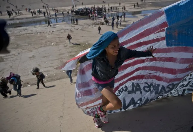 A migrant woman helps carry a handmade U.S. flag up the riverbank at the Mexico-U.S. border after getting past Mexican police at the Chaparral border crossing in Tijuana, Mexico, Sunday, November 25, 2018, as a group of migrants tries to reach the U.S. The mayor of Tijuana has declared a humanitarian crisis in his border city and says that he has asked the United Nations for aid to deal with the approximately 5,000 Central American migrants who have arrived in the city. (Photo by Ramon Espinosa/AP Photo)
