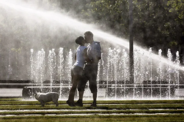 A couple kiss each other in front of water sprinklers as they walk the dog on a sunny morning near the Chancellery in Berlin, Wednesday, June 18, 2014. (Photo by Markus Schreiber/AP Photo)