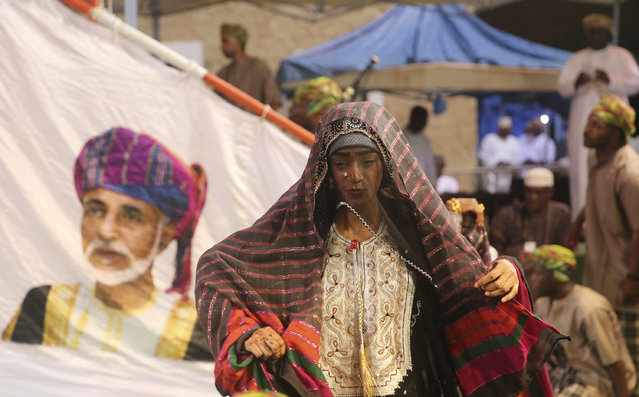 In this August 4, 2017 file photo, members of a dance troupe, in traditional garb, wait to perform next to a giant sail adorned with a portrait of the Sultan of Oman, Qaboos al-Said, during a festival celebrating the monsoon season, in Salalah, southern Oman. The 60-day festival of dance competitions, concerts, exorcisms performed by Sufis and the moderate temperatures draws tens of thousands of tourists to the region each year. (Photo by Sam McNeil/AP Photo)
