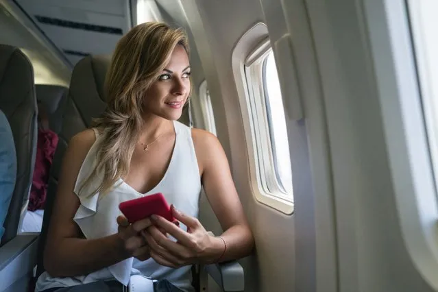 Beautiful woman traveling by plane and using her cell phone while looking through the window – lifestyle concepts (Photo by Hispanolistic/Getty Images)