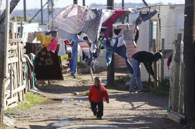 A child walk on a dirt road near his home as people nearby paint shacks with white fire retardant paint as part of their contribution to International Nelson Mandela day celebrating former South African president Mandela birth day in the township of  Nomzamo , South Africa,  Saturday, July 18, 2015. Thousands of South Africans celebrating former South African president Nelson Mandela birthday by giving 67 minutes of their time to help other people. (Photo by Schalk van Zuydam/AP Photo)