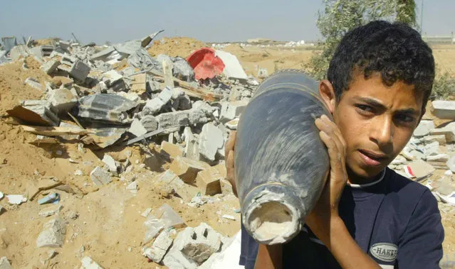 A Palestinian boy holds an Israeli artillery shell as he walks next to the remains of a house after it was demolished by Israeli troops before they pulled out from Rafah camp in the southern Gaza strip August 8, 2006. (Photo by Ibraheem Abu Mustafa/Reuters)