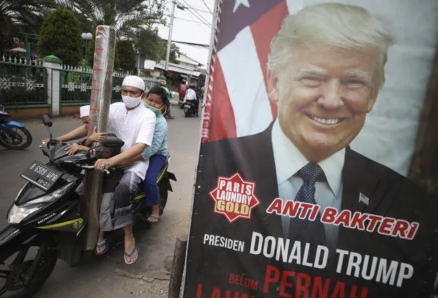 A man and his sons wearing face masks as a precaution against the coronavirus, ride past an advertisement using a portrait of U.S. President Donald Trump as a gimmick to attract people's attention in Ciputat, Indonesia, Friday, October 2, 2020. President Trump said early Friday that he and first lady Melania have tested positive for the coronavirus, a stunning announcement that plunges the country deeper into uncertainty just a month before the presidential election. Writings on the signboard read: “Anti-bacterial” and “President Donald Trump has never dropped his laundry here”. (Photo by Dita Alangkara/AP Photo)