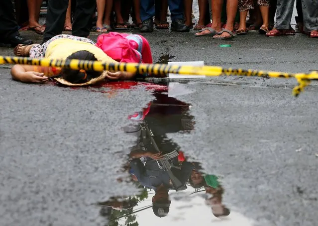 People and a policeman looking at the body of a woman, still clutching the school bag of her child, are reflected in a pool of water after she was shot while walking with her two children to school in Manila, Philippines, December 8, 2016. Police said the killing of the woman was the 13th recorded drug-related case in the past 24 hours in President Rodrigo Duterte's unrelenting war on drugs. (Photo by Bullit Marquez/AP Photo)