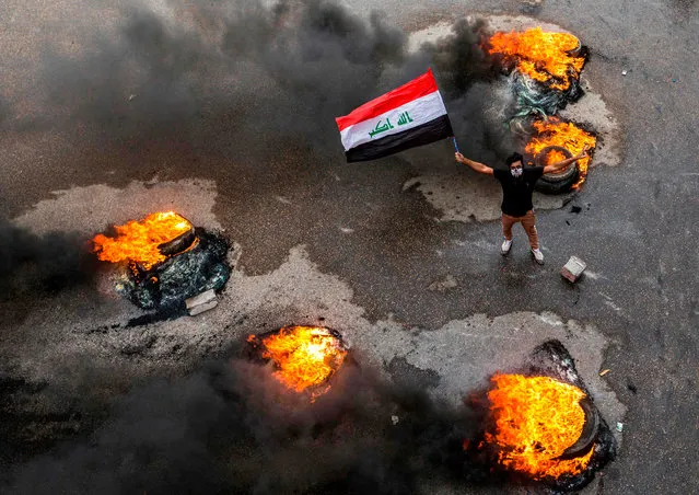 An Iraqi protester wearing a face bandana and waving a national flag poses past burning tires blocking a road during a demonstration in the southern city of Basra on November 17, 2019, as protesters cut-off roads and activists call for a general strike. Iraqis flooded the streets of the capital and southern cities in a general strike that bolstered the weeks-long movement demanding a government overhaul. Protesters cut roads in the oil-rich port city of Basra by burning tyres and in Hillah, south of Baghdad, students and other activists massed in front of the provincial headquarters. (Photo by Hussein Faleh/AFP Photo) 