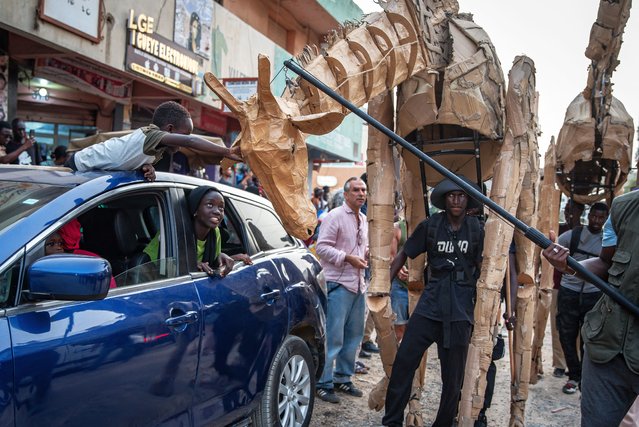 A child lying on a car roof touches a life size animal puppets moved by a puppeteer, part of the Herds public art and climate action project, in Ouakam district in Dakar on 26 April, 2025. A wildebeest, a gorilla and a giraffe are among the life-size puppets pursuing a 20,000-kilometre (12,400-mile) trek across the globe that started on April from the DR Congo capital, to raise awareness about migration due to climate change. The animals are part of The Herds project and aim to have crossed around 10 countries between central Africa and the Arctic by August. Their journey are taking them through Nigeria, Senegal, Morocco, France and Norway, among other countries. (Photo by Nicolas Remene/AFP Photo)