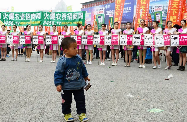 A child stands in front of models holding advertisement boards during a chemical fertilizer trade fair in Zhengzhou, Henan province, March 30, 2016. (Photo by Reuters/China Daily)