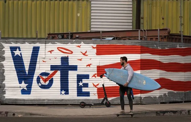 A man carries his surf board past a vote sign painted on a wall Monday, November 2, 2020, in the Venice Beach section of Los Angeles. (Photo by Jae C. Hong/AP Photo)
