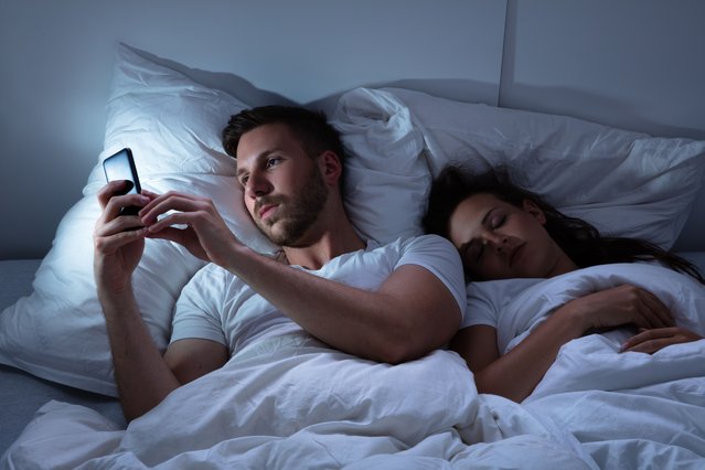 Young man using cellphone while her wife sitting on bed at night. (Photo  by Andrey Popov/Getty Images)