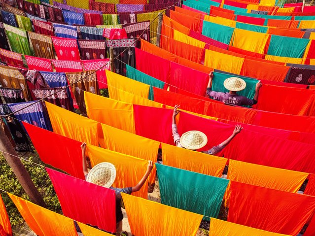 Workers hang hundreds of color-dyed sheets of cloth on a bamboo framework to dry in a dyeing factory in Narayanganj, Bangladesh on May 23, 2023. The drying process usually takes 4 hours, with each set of 200 pieces at a time to dry in temperatures over 42 degrees Celsius. Workers use hats for protection from the scorching heat because they have to constantly turn the colorful fabrics so that they dry perfectly in the sunlight. (Photo by Joy Saha/ZUMA Press Wire/Rex Features/Shutterstock)