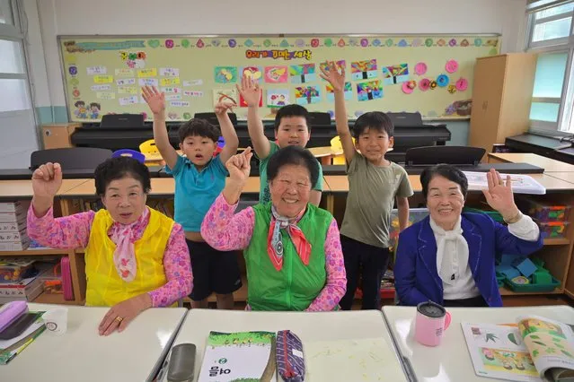 This picture taken on May 15, 2019 shows elderly South Korean women (front L-R) Park Young-ae, Nam Yang-soon and Shin Dong-hee posing and their eight-year-old classmates in a classroom at Woldeung Elementary School on the outskirts of Suncheon, 320 kilometres (198 miles) south of Seoul. With South Korea's population ageing rapidly and families migrating from the countryside to the cities for decades, rural primary schools are facing falling pupil numbers. Now some are targeting the opposite end of the age spectrum and recruiting elderly illiterate grandmothers – who were denied education on gender grounds during their own childhoods – to stave off the threat of closure and teach them to stave off the threat of closure and teach them to read and write. (Photo by Jung Yeon-Je/AFP Photo)