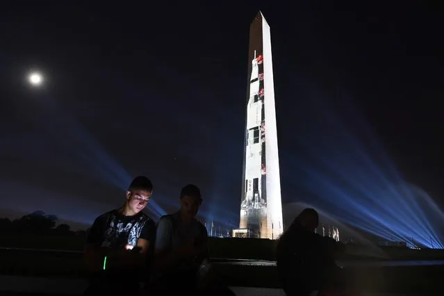 Tomas Mladenov is illuminated by his phone as an image of a Saturn V rocket is projected on the Washington Monument on July 16, 2019 to celebrate the 50th anniversary of the Apollo moon landing. (Photo by Matt McClain/The Washington Post)