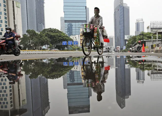 A drink vendor rides his bicycle reflected in a puddle in the main business district in Jakarta, Indonesia, Thursday, November 5, 2020. Indonesia's economy entered its first recession since the Asian financial crisis more than two decades ago as the country struggles to curb the coronavirus pandemic under control. (Photo by Dita Alangkara/AP Photo)