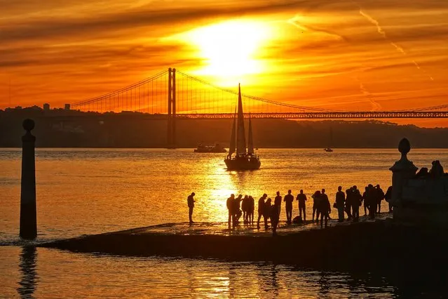 Locals and tourists alike stand on the banks of the Tagus River during sunset against the backdrop of the Ponte 25 de Abril (25th of April Bridge) suspension bridge at the Cais de Colunas, Portugal on January 1, 2022. (Photo by Soeren Stache/dpa-Zentralbild/dpa)