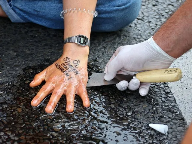 A police officer removes glue from the hand of a “Letzte Generation” (Last Generation) activist that reads “save oil instead of drill oil”, blocking a road under the slogan “Let's stop the fossil madness!” for an end to fossil fuels and against oil drilling in the North Sea, in Berlin, Germany, July 15, 2022. (Photo by Fabrizio Bensch/Reuters)